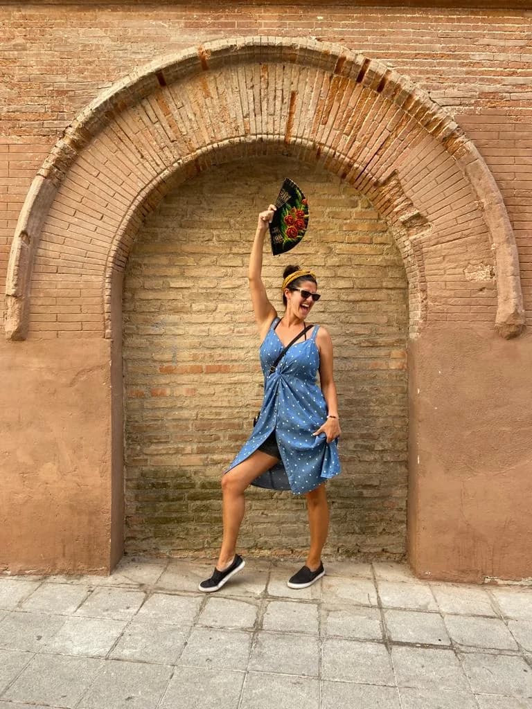 Annette Abreu smiling and holding a fan beneath a brick arch.
